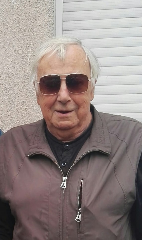 Starszy mężczyzna o siwych włosach pozuje z uśmiechem do zdjęcia. Na nosie ma kanciaste okulary przeciwsłoneczne.