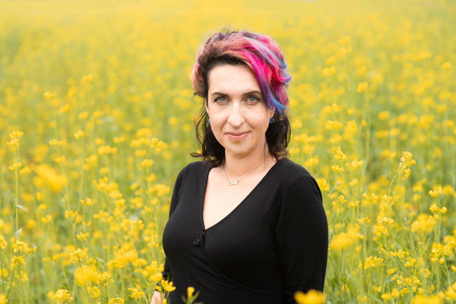 Uśmiechnięta kobieta stoi na polu wysokich żółtych kwiatów. Jej włosy są pomalowane na różne kolory.