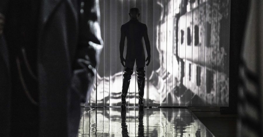 Scena spektaklu, odwrócony mężczyzna w czarnym kostiumie stoi naprzeciwko ściany, na której jest wyświetlony tunel. Na pierwszym stoi mężczyzna w długim płaszczu.
