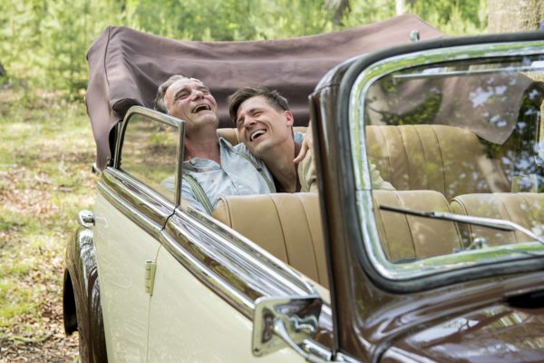 Fotos z filmu „Szarlatan”. Dwójka mężczyzn w średnim wieku, na tylnym siedzeniu eleganckiego kabrioletu, śmieje się jednocześnie tuląc się o siebie.