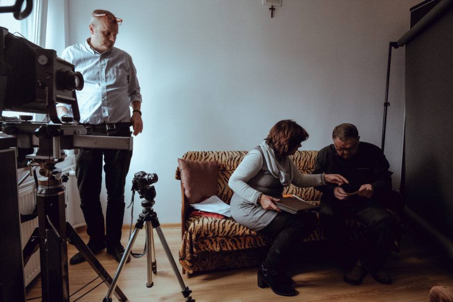 Kulisy powstawania filmu. Kobieta i mężczyzna siedzą na kanapie i przeglądają zdjęcia z albumu. Z boku przygląda im się mężczyzna, stojący nieopodal kamer filmowych na statywach.