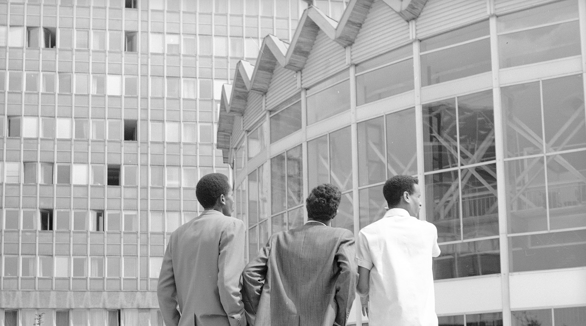 Trójka elegancko ubranych mężczyzn stojących przed Rotundą – przeszklonym budynkiem w kształcie walca.