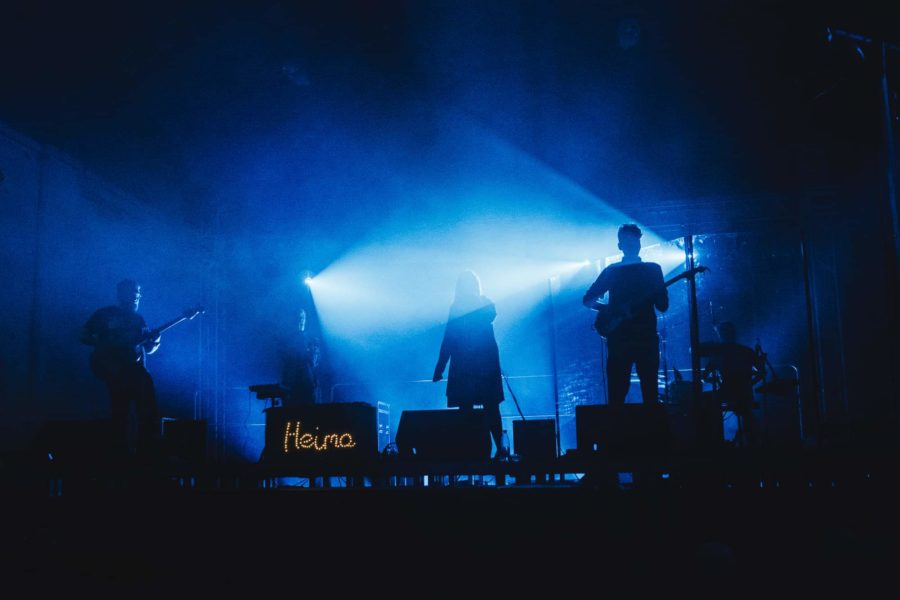 Zespół muzczny gra koncert. Dym na scenie i słabe światło sprawiają, że widać jedynie ciemne sylwetki członków zespołu.