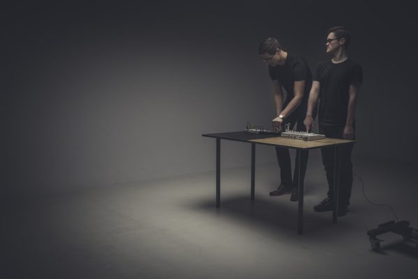 Dwóch mężczyzn tworzy muzykę elektroniczną na mikserach, ustawionych na ławce, w kącie sali.