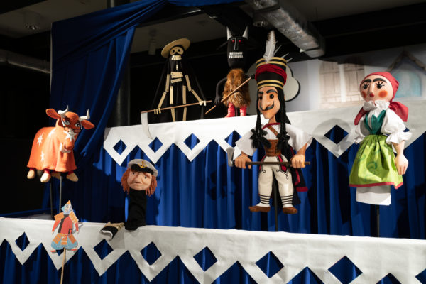 Dwie sceny teatru lalek, na bliższej pięć lalek przymocowanych prętami, na dalszej scenie dwie lalki przymocowane prętami.