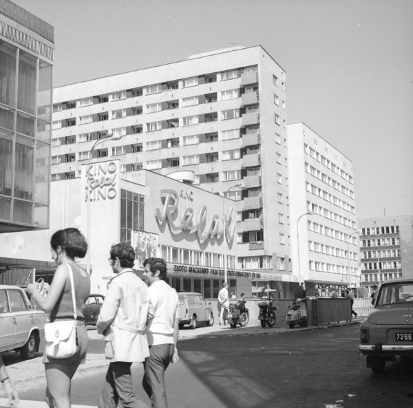 Trójka ludzi przechodzących przez ulicę w centrum miasta, na tle budynku z neonem Kino Relax