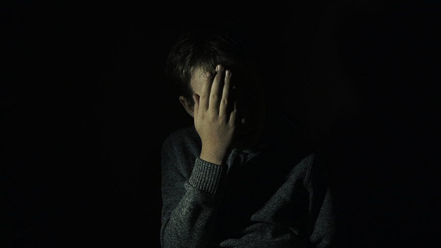 Częściowo oświetlony chłopczyk, zasłania ręką część twarzy. Chłopczyk znajduje się na ciemnym tle.