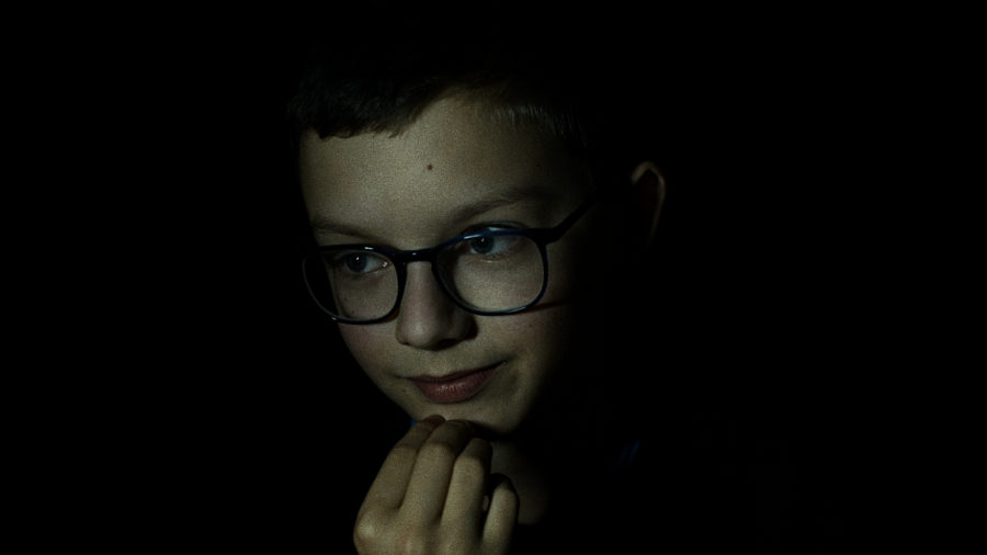 Chłopczyk w okularach nad czymś się zastanawia, drapie się ręką po brodzie. Postać znajduje się na ciemnym tle.