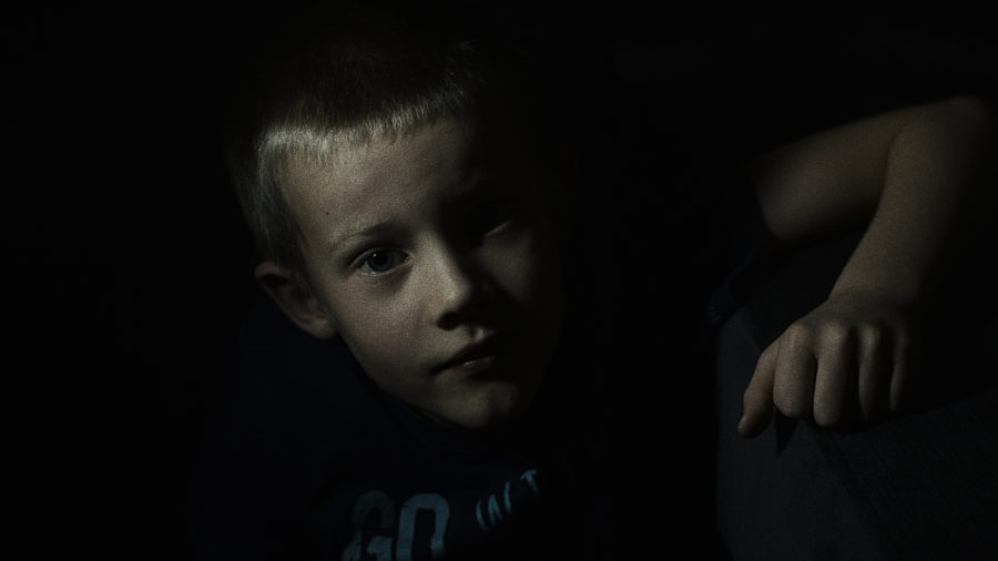 Częściowo oświetlony chłopczyk patrzy się w obiektyw, swoją prawą rękę opiera na szarym prostokątnym kształcie. Tło jest ciemne.