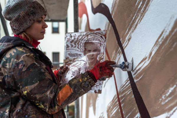 Kobieta na rusztowaniu, w jednej ręce trzyma projekt muralu, drugą ręką maluje.