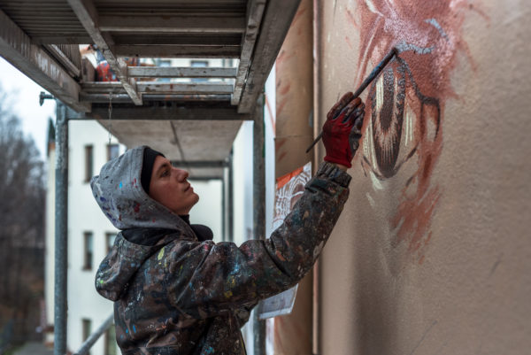 Mężczyzna stoi na rusztowaniu i pędzlem maluje mural.
