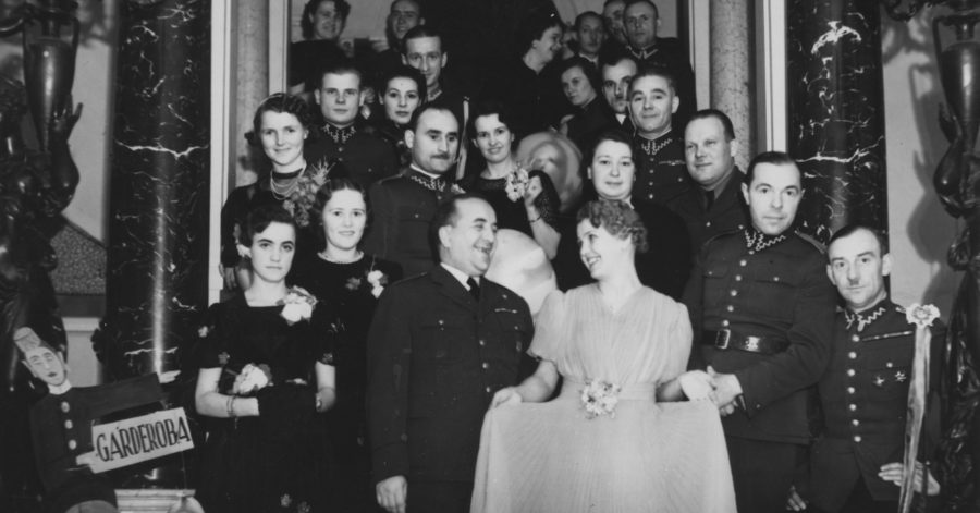 Czarnobiałą fotografia przedstawia, grupę elegancko ubranych ludzi, którzy pozują do zdjęcia w eleganckiej sali.
