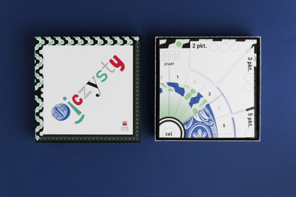 Górna część pudełka gry leży obok zwiniętej planszy do gry. Okładka gry przedstawia napis: "ojczysty", każda litera jest napisana innym fontem i ma różny kolor.